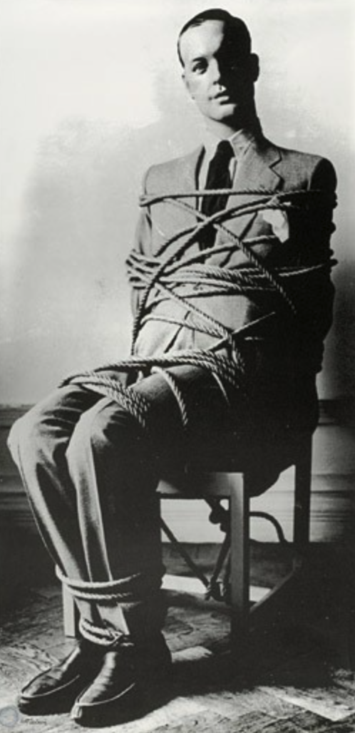 A man dressed in a suit sitting on a chair, bound with ropes across his body and legs.