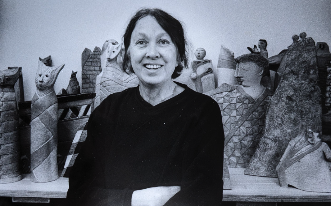 A black and white photo of a smiling woman standing in front of various ceramic sculptures and artworks.