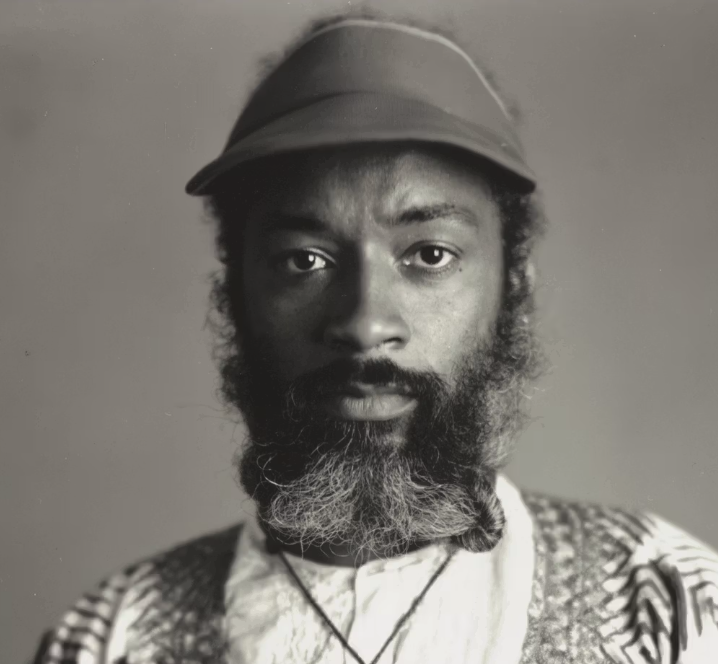 Black and white portrait of a man with a beard, wearing a cap and an ornate shirt, looking directly at the camera.