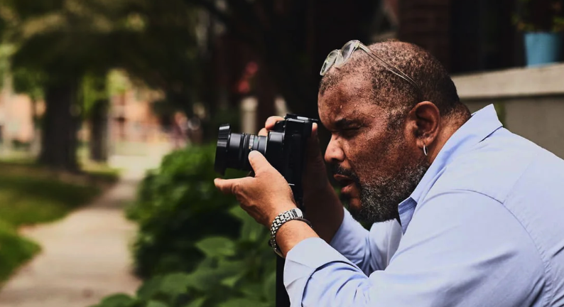 A man focused on taking a photograph with a camera, surrounded by greenery.
