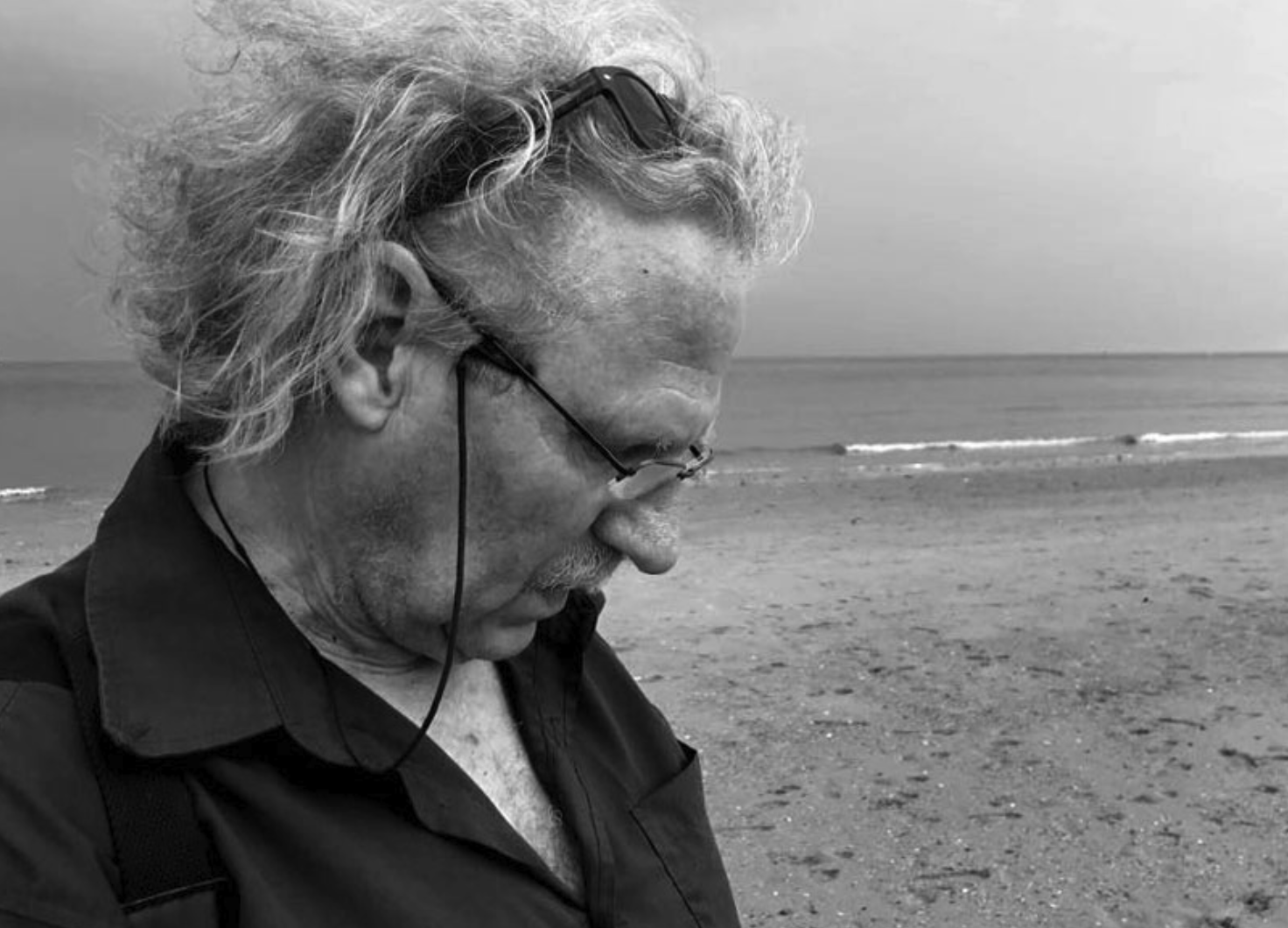 A man with curly hair and glasses stands on a beach, looking down. The image is in black and white, with a calm sea and cloudy sky in the background.