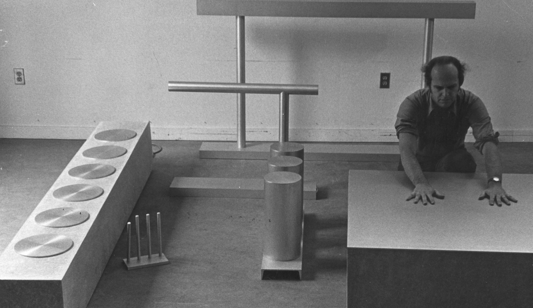 A black and white photograph of a man interacting with abstract geometric sculptures in a studio setting. The man is kneeling, with his hands on a flat surface, surrounded by various metal shapes including cylinders and a bar.