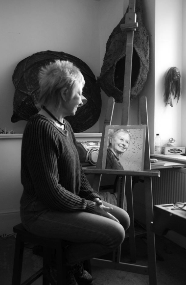 A woman with short, spiky hair sitting on a stool in an art studio, looking at a portrait of herself on an easel in front of her.