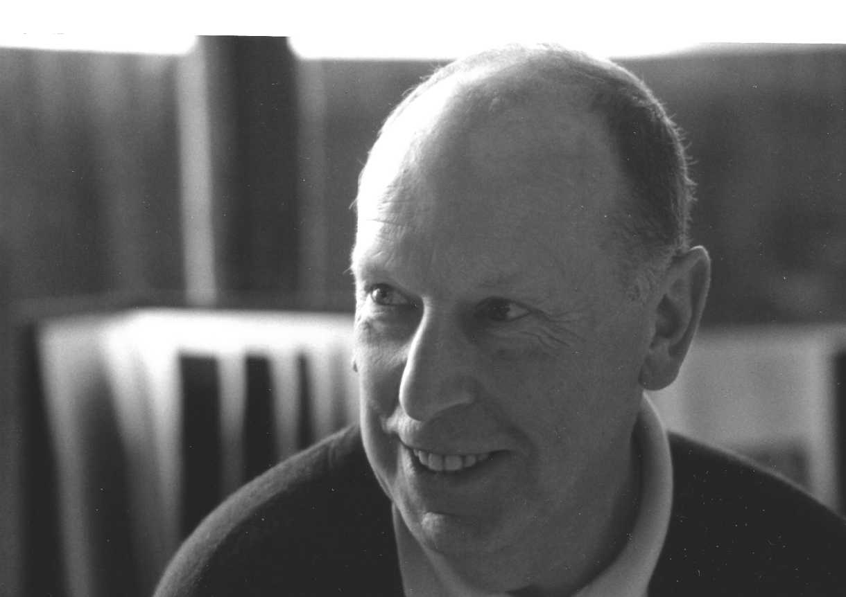 Black and white portrait of a smiling elderly man with light hair and a round face, wearing a dark sweater over a collared shirt, in a studio setting.