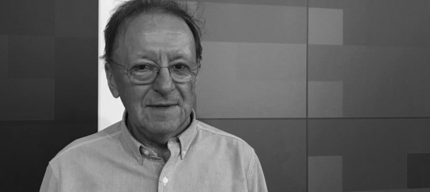 A man with glasses is smiling while standing against a textured wall.