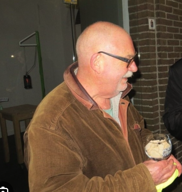 A bald man with glasses smiles while holding a glass with a light-colored beverage. He is wearing a brown jacket with a light-colored shirt underneath, standing in an indoor setting with a brick wall in the background.