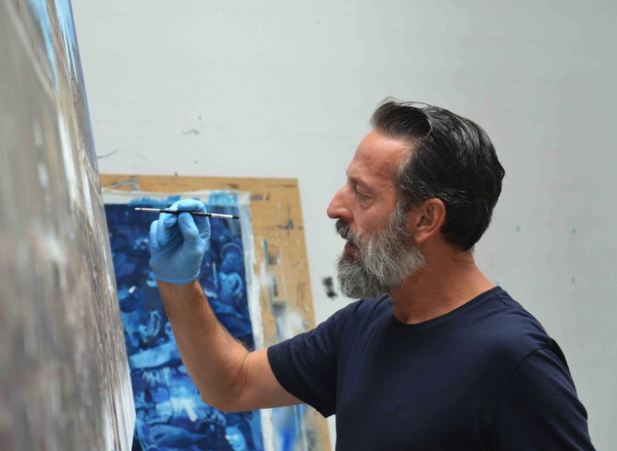 An artist wearing blue gloves is painting on a large canvas in a studio setting.