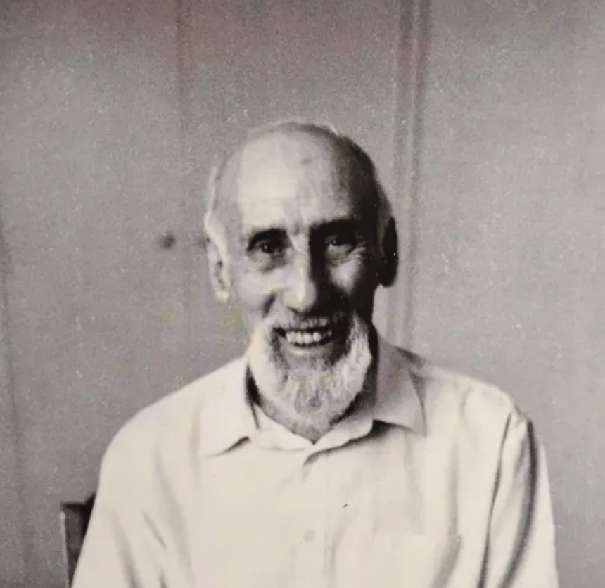 A smiling elderly man with a beard, wearing a light-colored shirt, seated in a simple interior setting.
