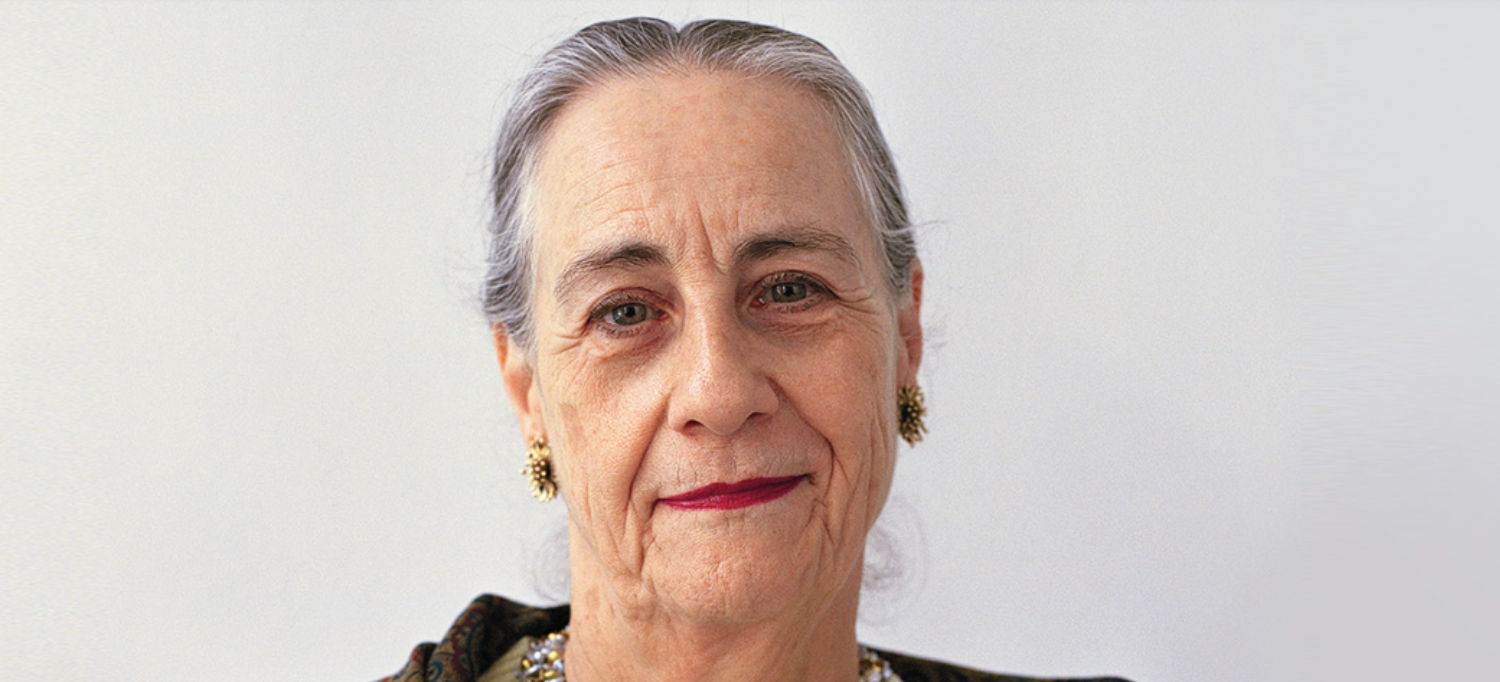 A close-up portrait of an elderly woman with gray hair, wearing elegant earrings and lipstick, looking directly at the camera against a plain background.