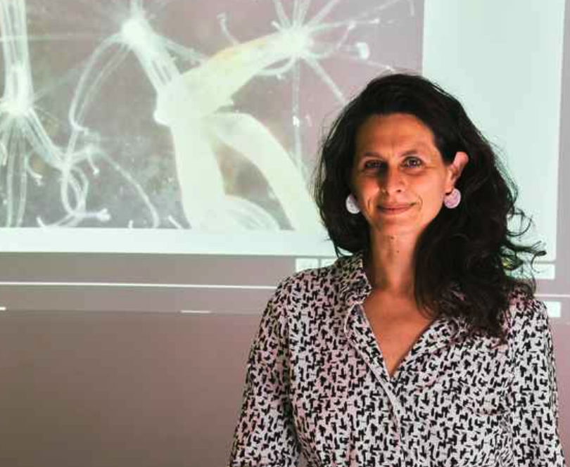 A woman with long, dark hair stands in front of a projected image of sea creatures, smiling. She is wearing a patterned shirt and large earrings.