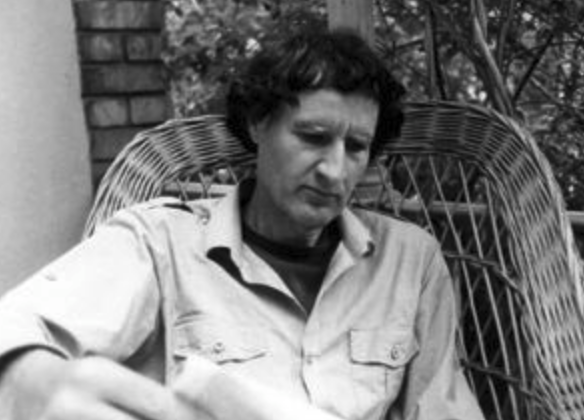 A man with wavy dark hair sits in a wicker chair, looking down thoughtfully while holding a piece of paper.