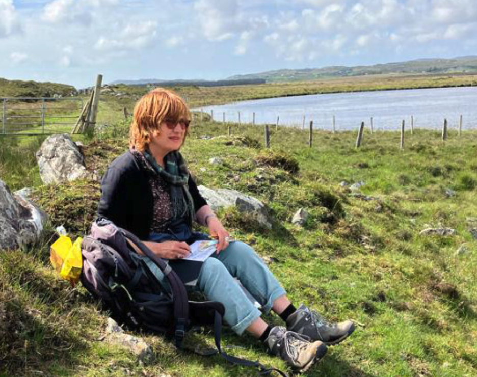 A person sitting on grassy ground near a water body, wearing casual clothes and boots, with a backpack and notebook in hand, enjoying a sunny day outdoors.