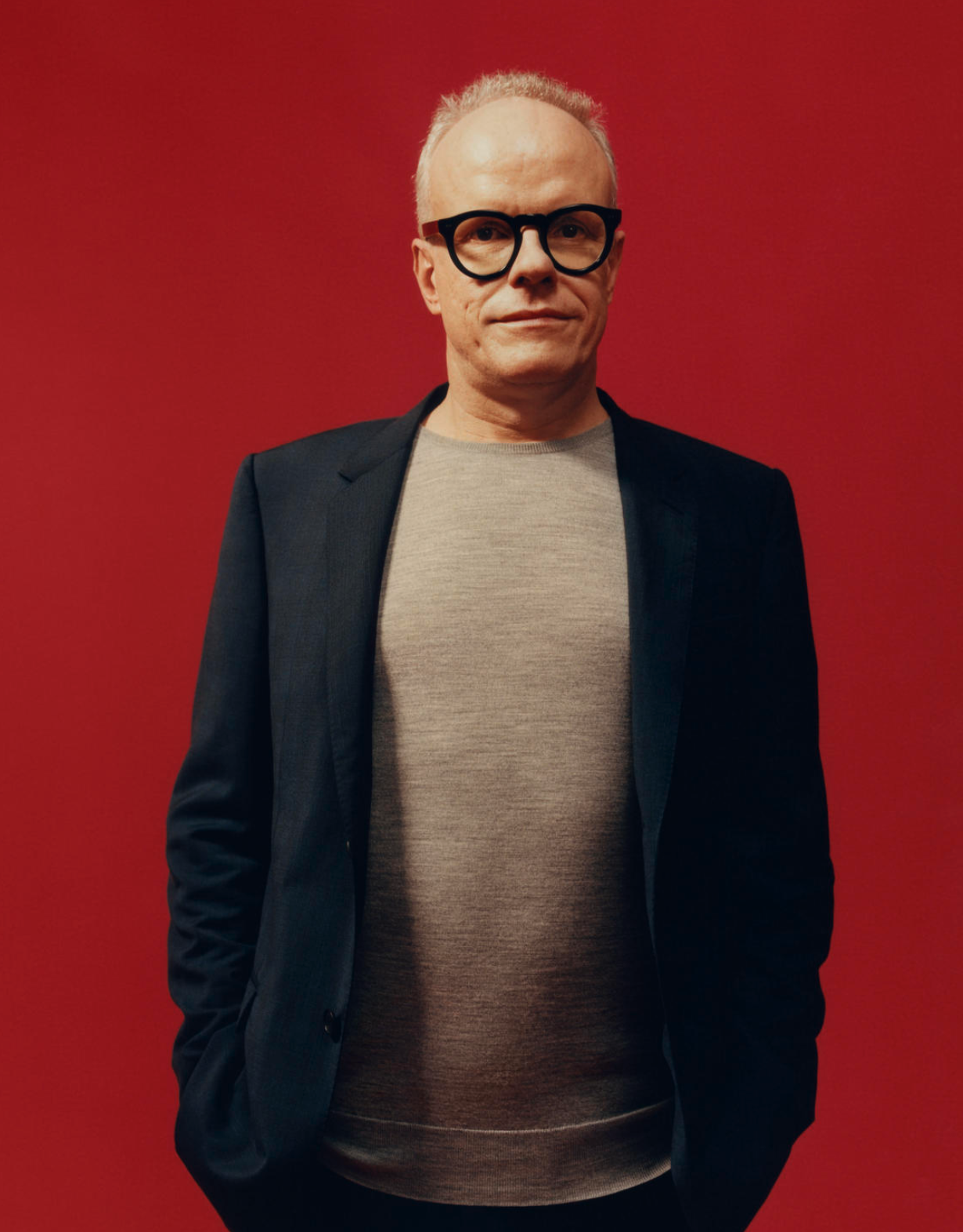 A man wearing large black glasses and a dark blazer over a gray sweater stands against a red background.