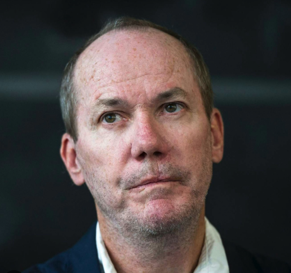 Close-up of a man with a serious expression, wearing a light shirt and dark jacket, against a blurred dark background.