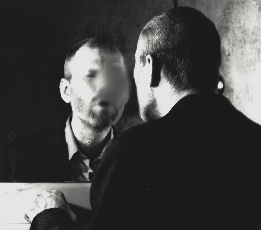A black and white photograph of a man looking into a mirror, with his face obscured, symbolizing themes of identity and reflection.