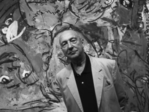 A man in a light-colored blazer stands in front of a colorful abstract painting, featuring various shapes and figures.