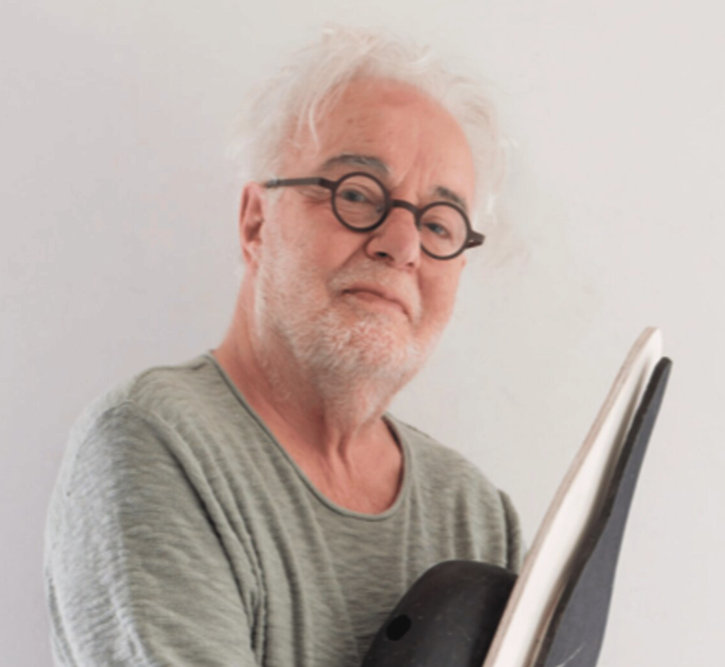 A senior man with white hair and glasses holding a black object while posing against a plain white background.